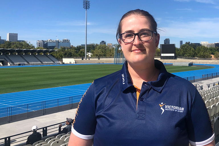 A woman, wearing a blue shirt with text on the shirt reading Victorian Institiute of Sport, standing with a grass oval and city skyline in the background. 