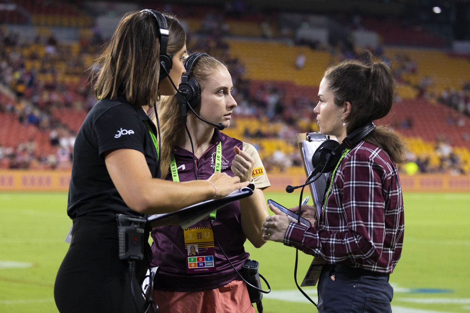 Three women on a sports field wearing communication headsets  and carrying clipboards.
