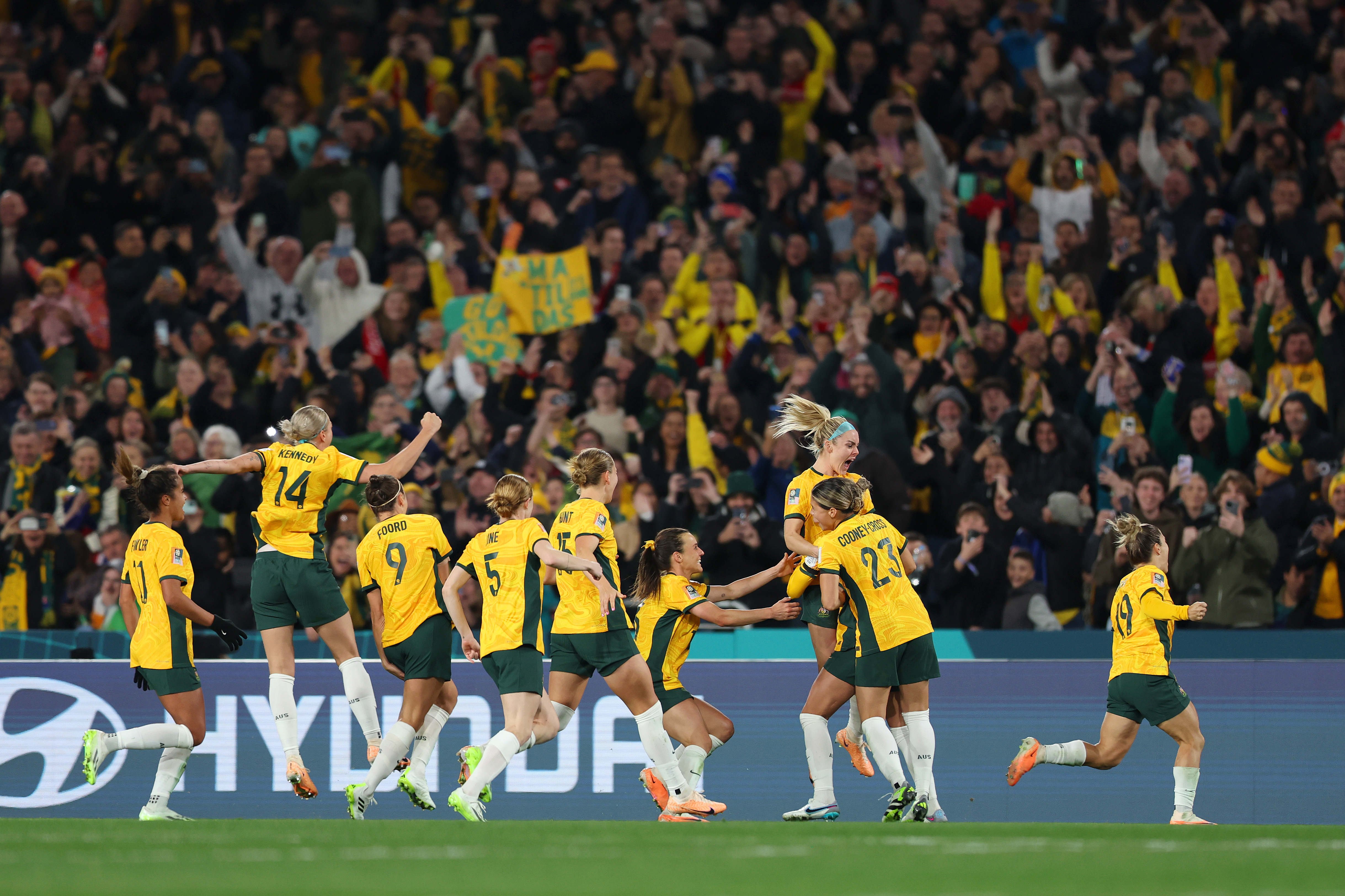A group of Matildas players run together to celebrate on the field, with fans cheering from the stands.