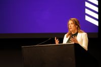 A woman stands behind a lectern, in front of a blue screen